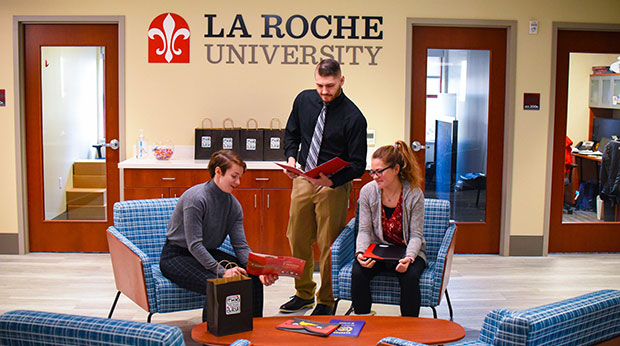 Two Admissions Counselors talk to a student in the La Roche University Welcome Center. One counselor and the student are seated, and one counselor is standing. All three of them look at brochure together. The La Roche University logo is behind them on the wall. 