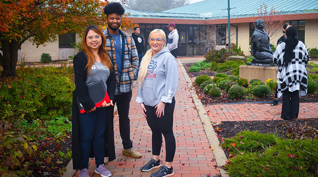 Three La Roche University students stand on the walkway outside of Wright Library. Two students are behind them, and two students stand to the right of them, talking.