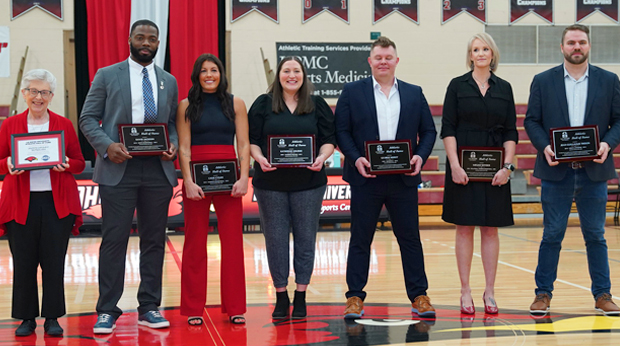 Seven La Roche University alumni athletes hold plaques and stand in the middle of the basketball court of the Kerr Fitness & Sports Center at an Athletic Hall of Fame induction ceremony. 