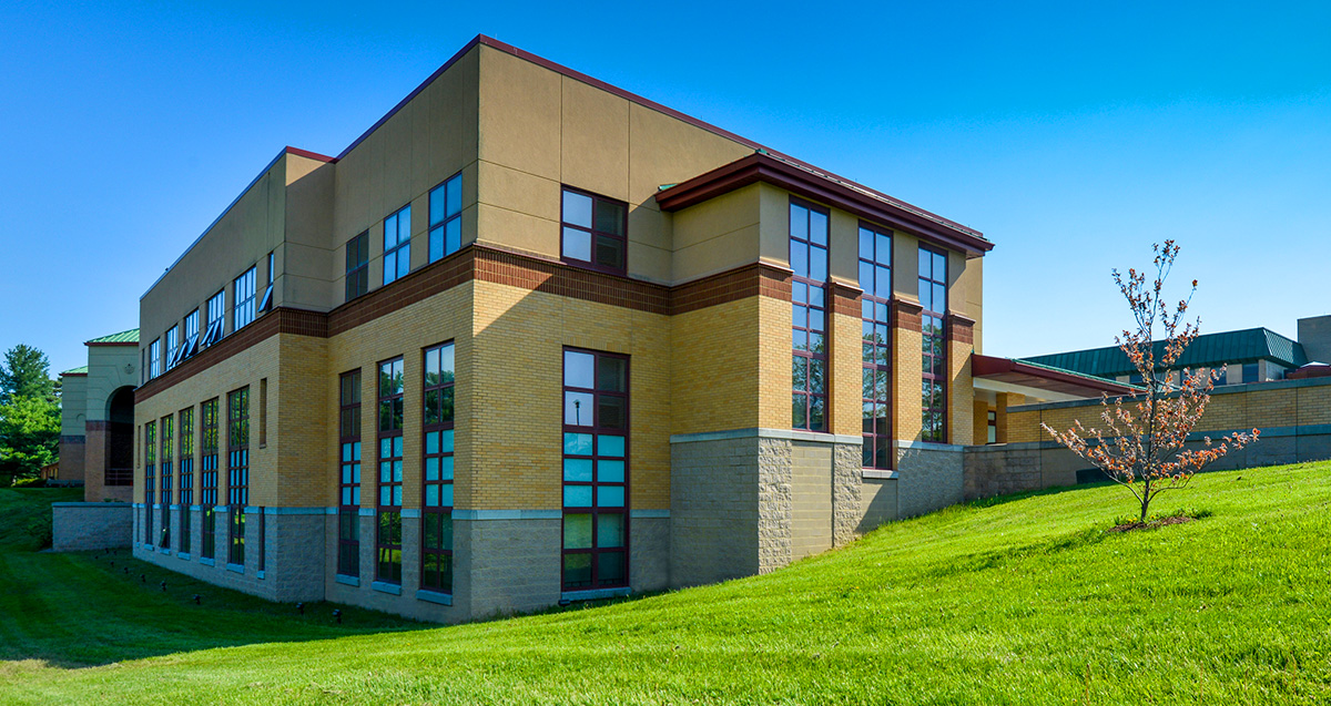 Exterior view of La Roche University's Huber Academic Center building with yellow and beige walls, large windows, and surrounded by green lawns under a clear blue sky.