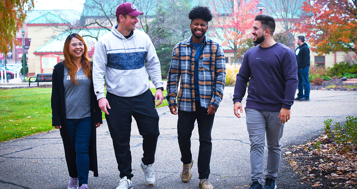 Four college students walking together on a campus path in autumn, smiling and talking.