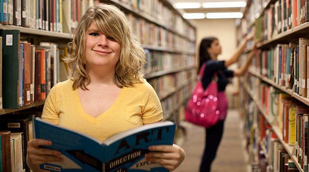 A La Roche University student stands in an aisle of the Wright Library. She is holding an open book, and a student with a bright pink purse is browsing the collection behind her in the background.