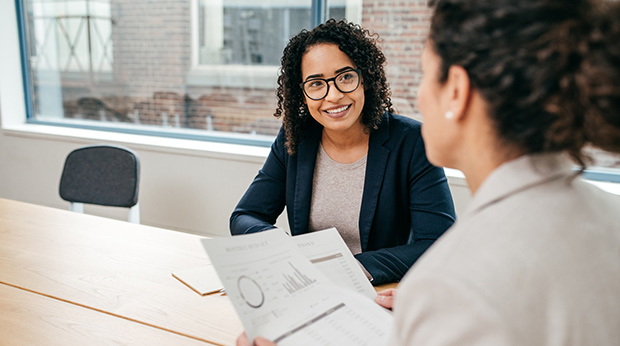 Two people sit at a conference table. One is holding spreadsheets.
