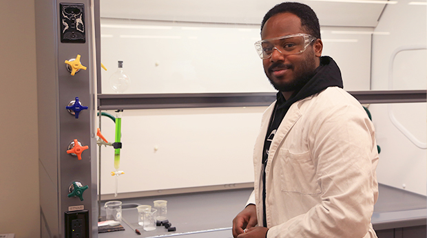 A La Roche University student wearing a lab coat and protective eyewear stands in front of equipment in a chemistry lab.