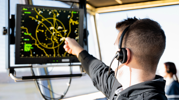 A male student pointing at the air traffic control radar screen. He is wearing a headset and sitting in an air traffic control tower. There is a female student wearing a headset in the background.