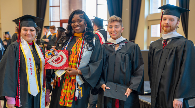 Four graduates in La Roche University's Zappala Campus Center Square building. They all wear their graduation gowns, and two are wearing their caps. One cap is decorated.