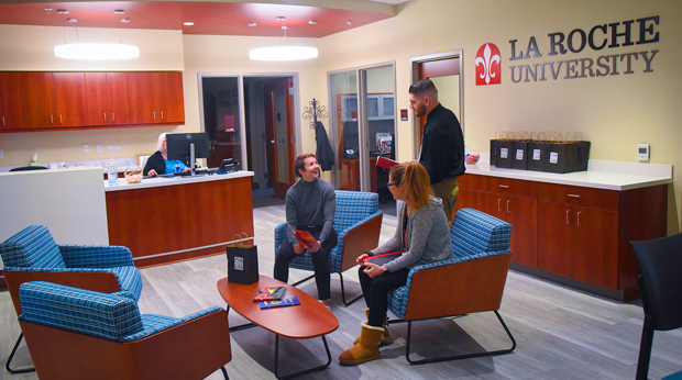 Two admissions counselors and a student sit and talk to each other in the Welcome Center at La Roche University.  The University logo is on a wall behind them. A staff member works at a computer in th