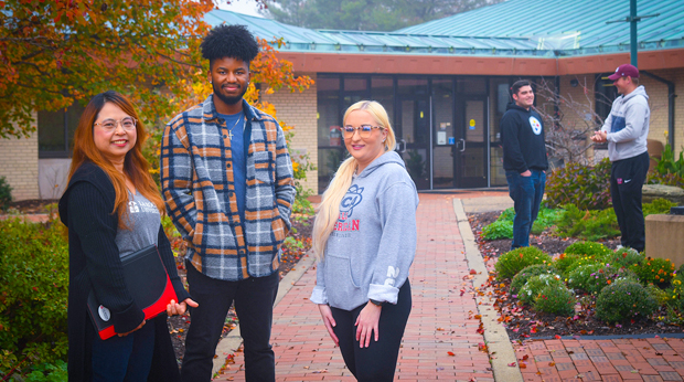Three La Roche University students stand on the walkway in front of Wright Library. Two students converse in the background behind them.