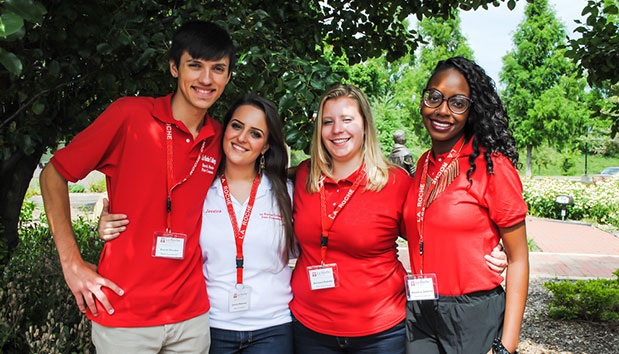 Four La Roche University students stand outside in front of a tree. One is wearing a white shirt, and the others are wearing red shirts. All of the students have lanyards around their necks. 