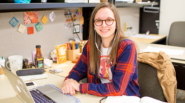 A La Roche University student sits at a desk with their open laptop and an open book beside them. She is smiling at the camera.