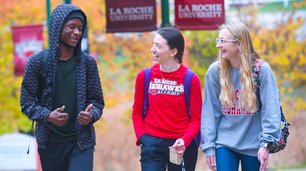 Three students walking and chatting on the La Roche University campus, with one wearing a La Roche University Redhawks sweatshirt. Autumn leaves visible in the background.  