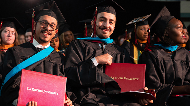 Two La Roche University graduates hold their diplomas at commencement. They sit with a crowd of other students wearing caps and gowns.
