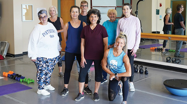 A group of Adventures in Lifelong Learning members at La Roche University stand in the fitness studio of the Kerr Fitness & Sports Center. There are dumbbells near them on the floor, and a mirror is b
