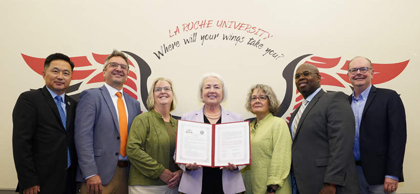 Group of seven individuals standing side by side with the La Roche University “Where will your wings take you?” wings graphic in the background. One person is displaying a certificate towards the camera. They are dressed in business attire.