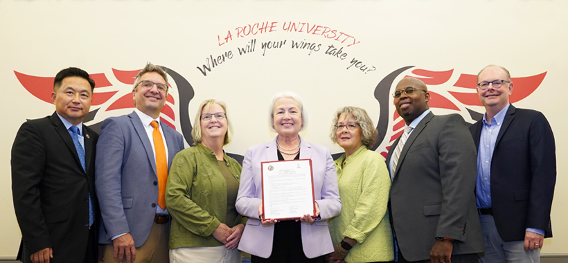 Group of seven individuals standing side by side with the La Roche University “Where will your wings take you?” wings graphic in the background. One person is displaying a certificate towards the camera. They are dressed in business attire.