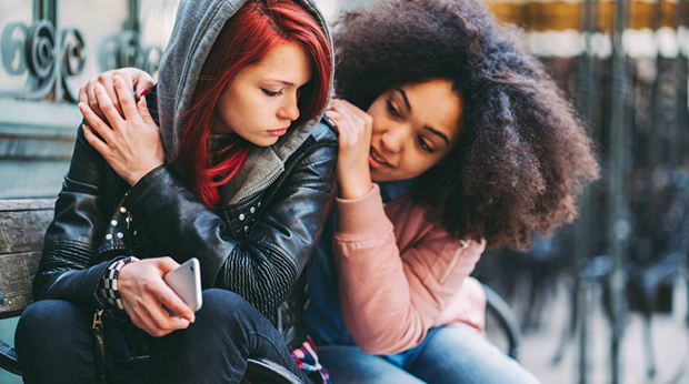 Two people sit on the ground outside hugging each other.